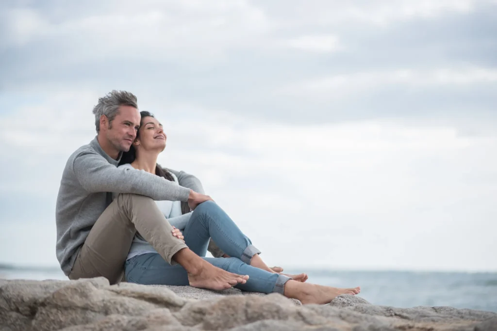 A couple sitting barefoot on a rocky beach, leaning against each other and enjoying a peaceful moment by the ocean under a cloudy sky.